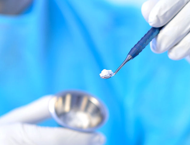 Gloved hands lifting granular bone graft material on a small dental instrument above a metal dish